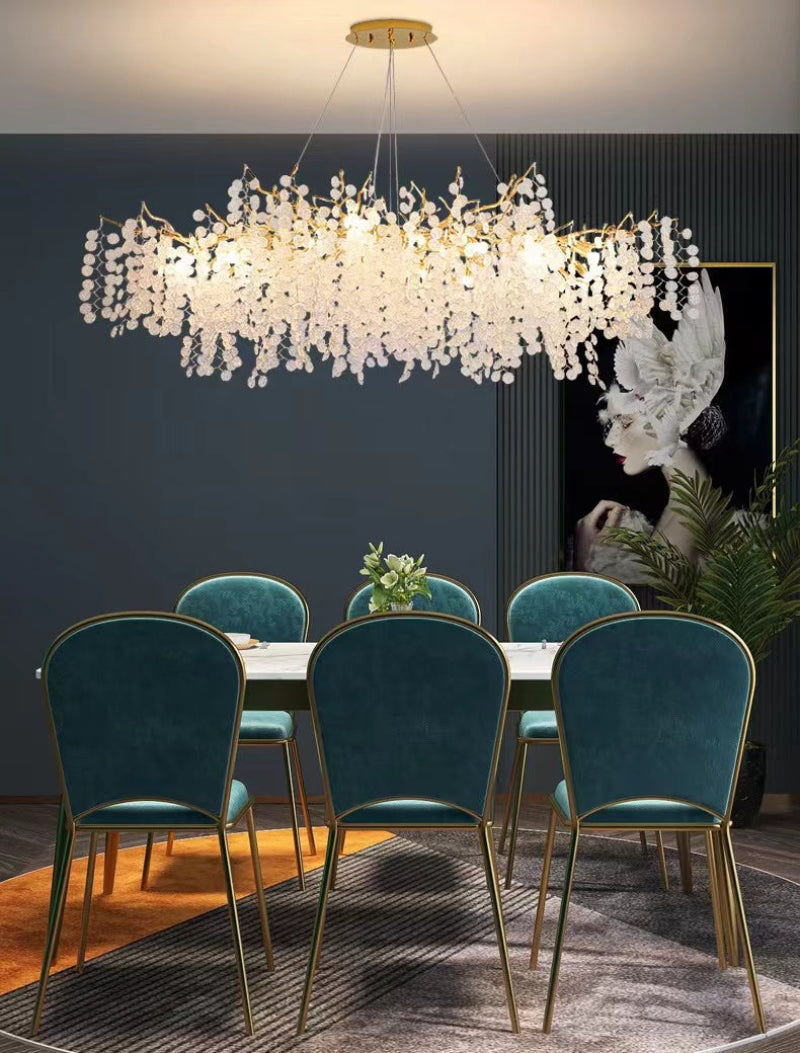 A modern linear tree branch chandelier with glass-like leaves hanging over a dining table with blue chairs, providing a warm white light.