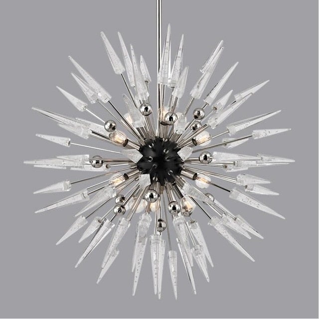 A unique sputnik chandelier with a gold or light grey finish, featuring a round, star-like shape with multiple arms, hanging over a dining table with chairs around it, set against a dark marble wall.